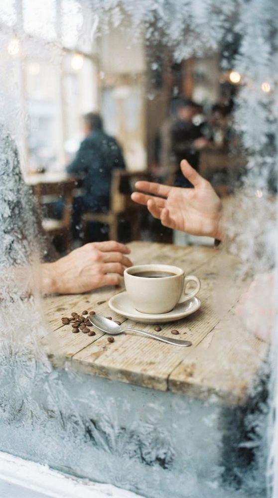 Mãos a conversar num café através de janela enevoada com chávena de café e grãos na mesa