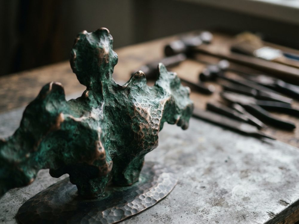 Escultura abstrata de bronze texturizado sobre mesa com ferramentas ao fundo