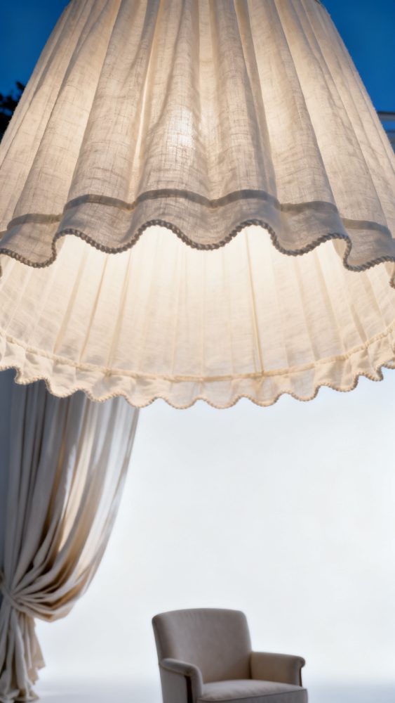 Candeeiro pendente branco com bordas onduladas, cortina clara e poltrona cinza clara num ambiente calmo