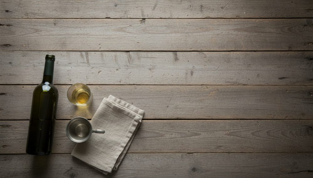 Garrafa de vinho branco, copo cheio, pequeno jarro metálico e pano sobre mesa de madeira clara