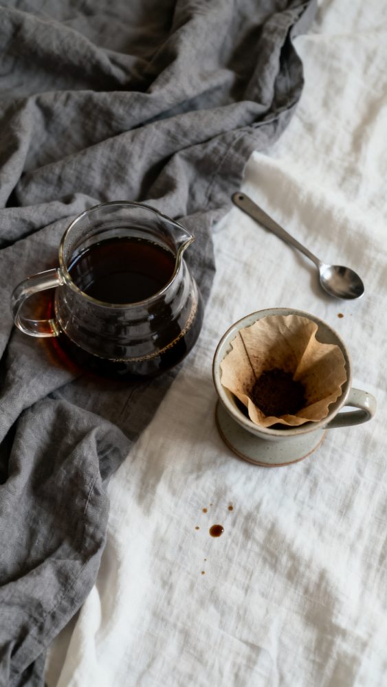 Jarra de vidro com café coado, caneca com filtro de papel e colher sobre tecido branco e cinzento