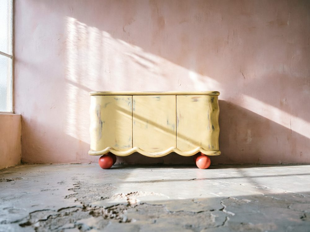 Aparador amarelo vintage com pés vermelhos numa sala com parede rosa e chão rugoso