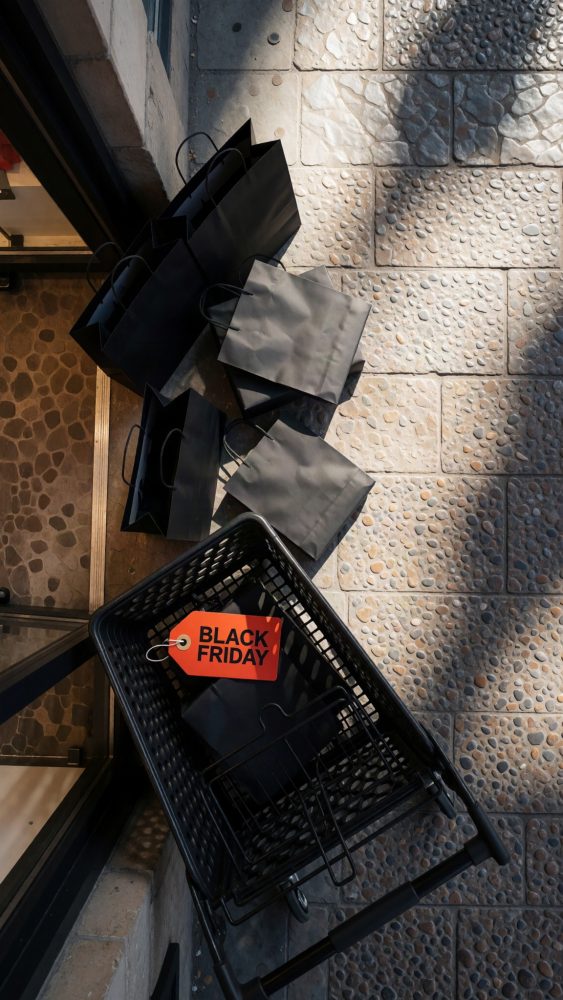 Carrinho de compras preto com sacos pretos e etiqueta Black Friday vermelha sobre chão de pedra.