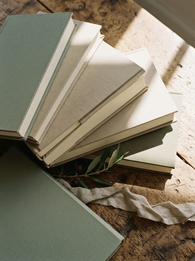 Seis livros com capas de tecido bege e verde empilhados sobre mesa de madeira rústica iluminada naturalmente