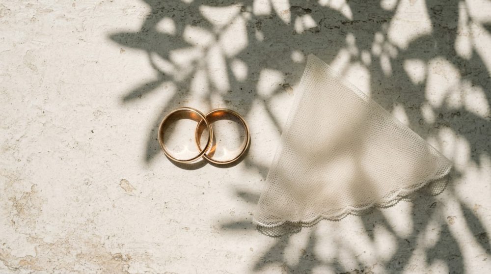 Duas alianças douradas ao lado de véu de tule sob sombras de folhas
