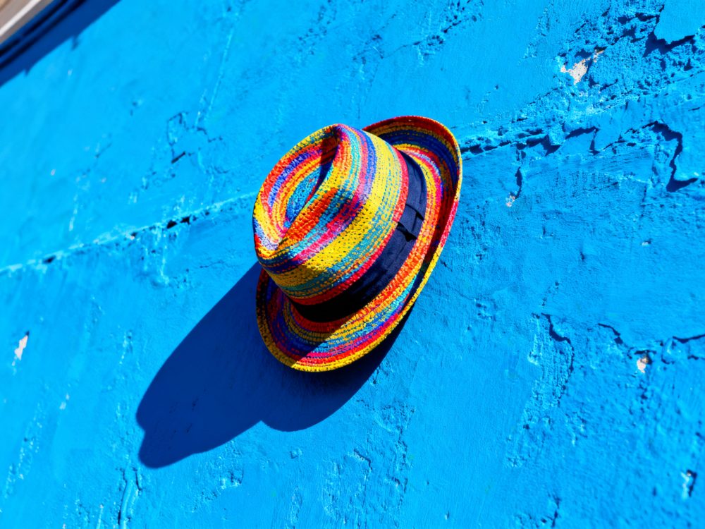 Chapéu de palha às riscas multicoloridas sobre parede azul texturada, com sombra longa.