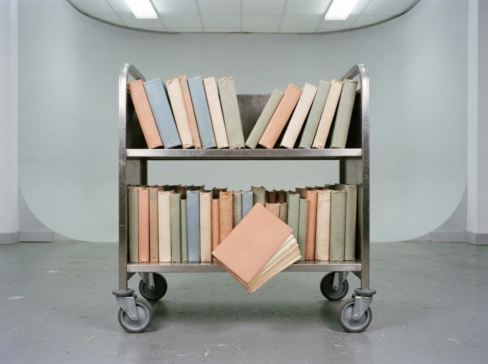 Carrinho metálico de biblioteca com livros de capas pastel e um livro aberto suspenso na prateleira inferior.