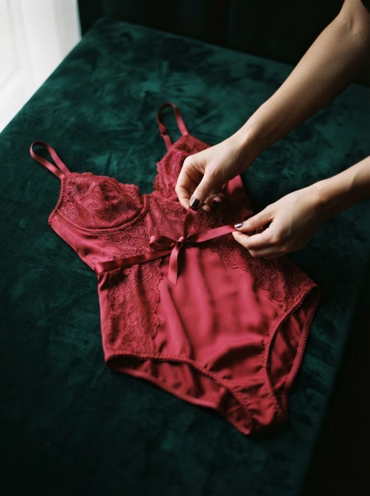 Body grená em cetim e renda floral com laço à cintura sobre veludo verde-escuro, ajustado por mãos femininas.