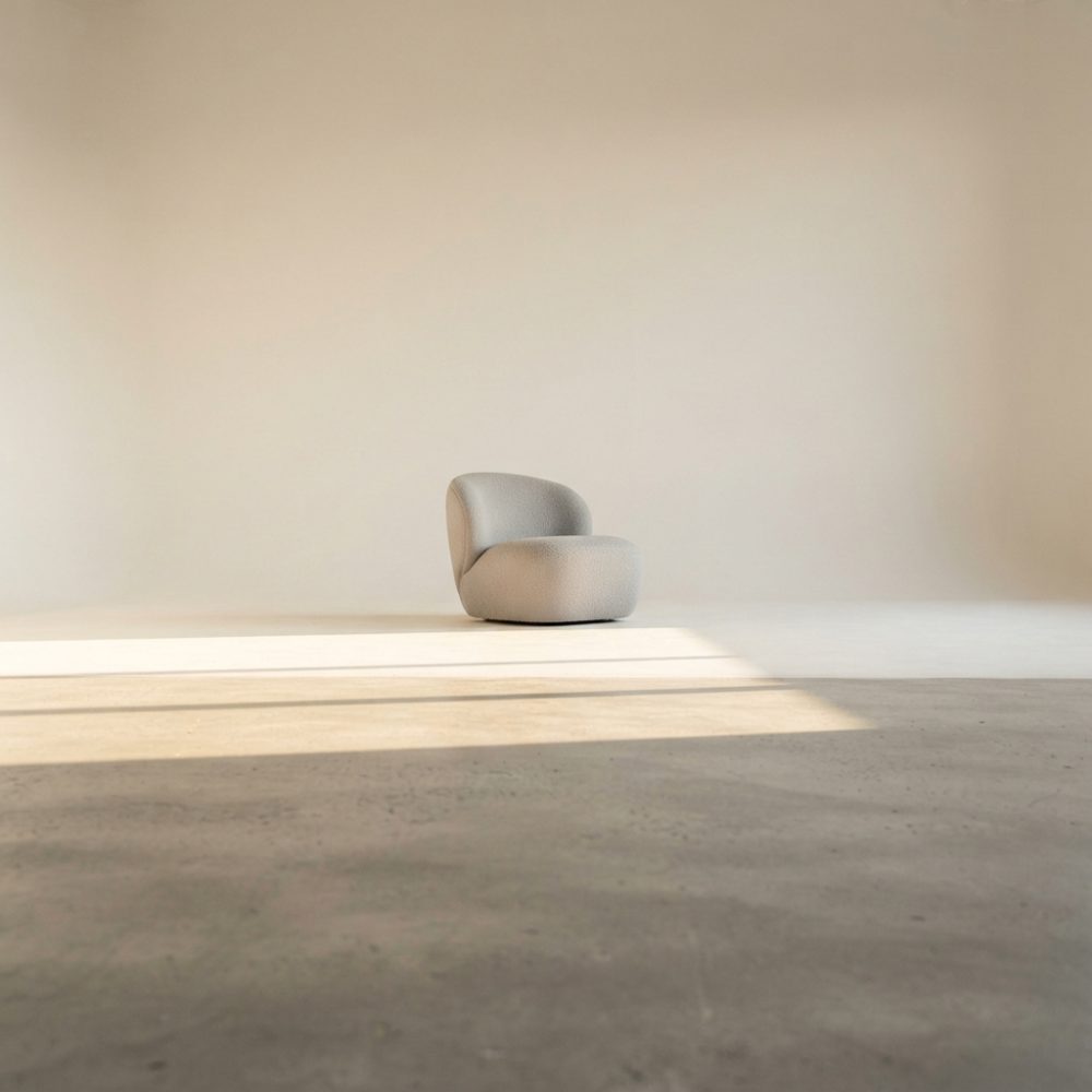 Poltrona curva bege num interior minimalista de paredes claras e chão em betão, iluminado pelo sol.