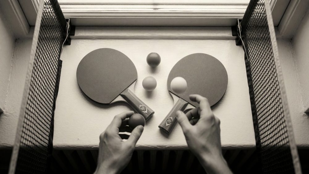 Duas raquetes de ténis de mesa, bolas e mãos sobre um parapeito branco entre grades metálicas.