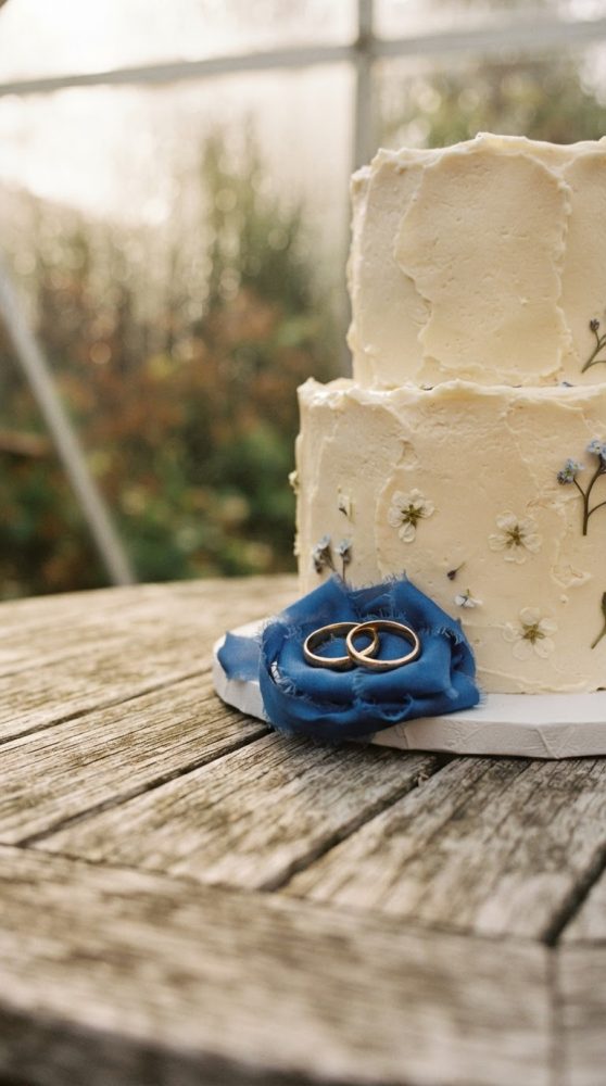 Bolo de casamento creme com alianças douradas sobre flor de tecido azul numa mesa de madeira