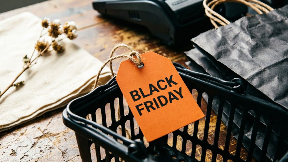 Cesto de compras preto com etiqueta laranja Black Friday sobre mesa de madeira, sacos escuros e terminal de pagamento.