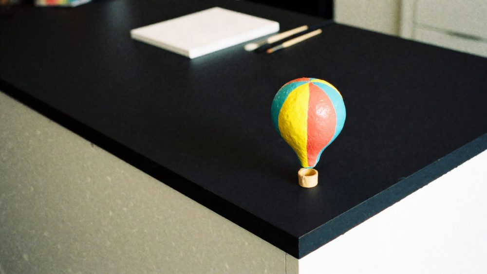 Balão de ar quente em miniatura amarelo, coral e azul sobre mesa preta com pincéis e bloco de papel.