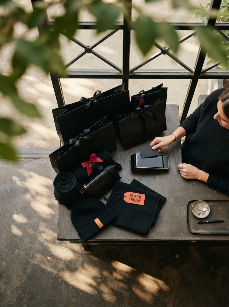 Sacos de compras pretos, roupa e etiqueta Black Friday sobre mesa rústica, junto a uma mulher com telemóvel.