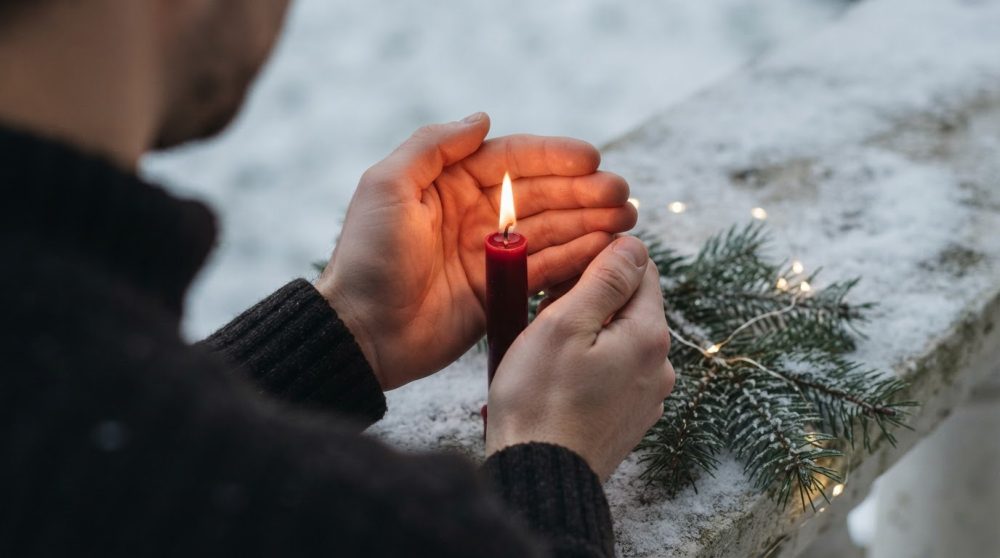 Mãos protegem uma vela vermelha acesa junto a ramos de pinheiro com neve e luzes de Natal.