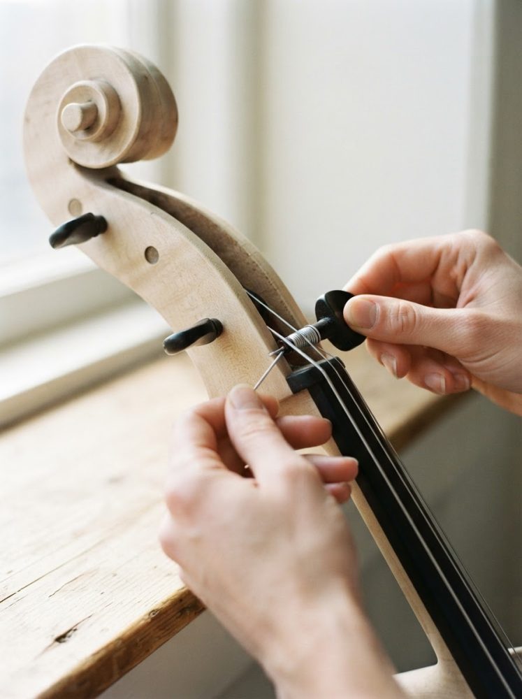 Mãos a ajustar as cordas de um violoncelo de madeira clara junto a uma janela