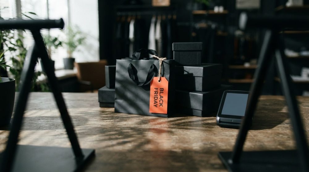 Saco preto e caixas empilhadas com etiqueta laranja Black Friday sobre mesa de madeira