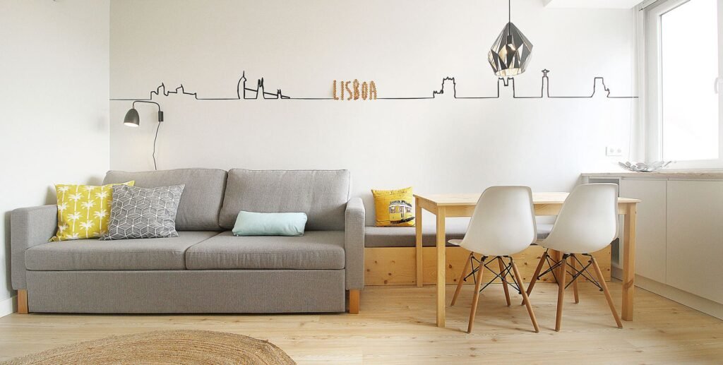 Sala com sofá cinzento, almofadas coloridas e mesa de refeições em madeira, com a silhueta de Lisboa desenhada na parede