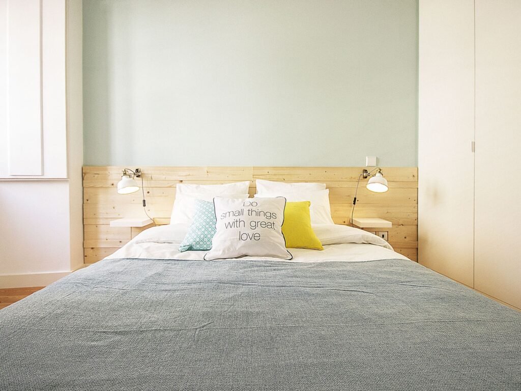 Quarto com cabeceira em madeira clara, colcha cinzenta e almofada branca com a frase «Do small things with great love»