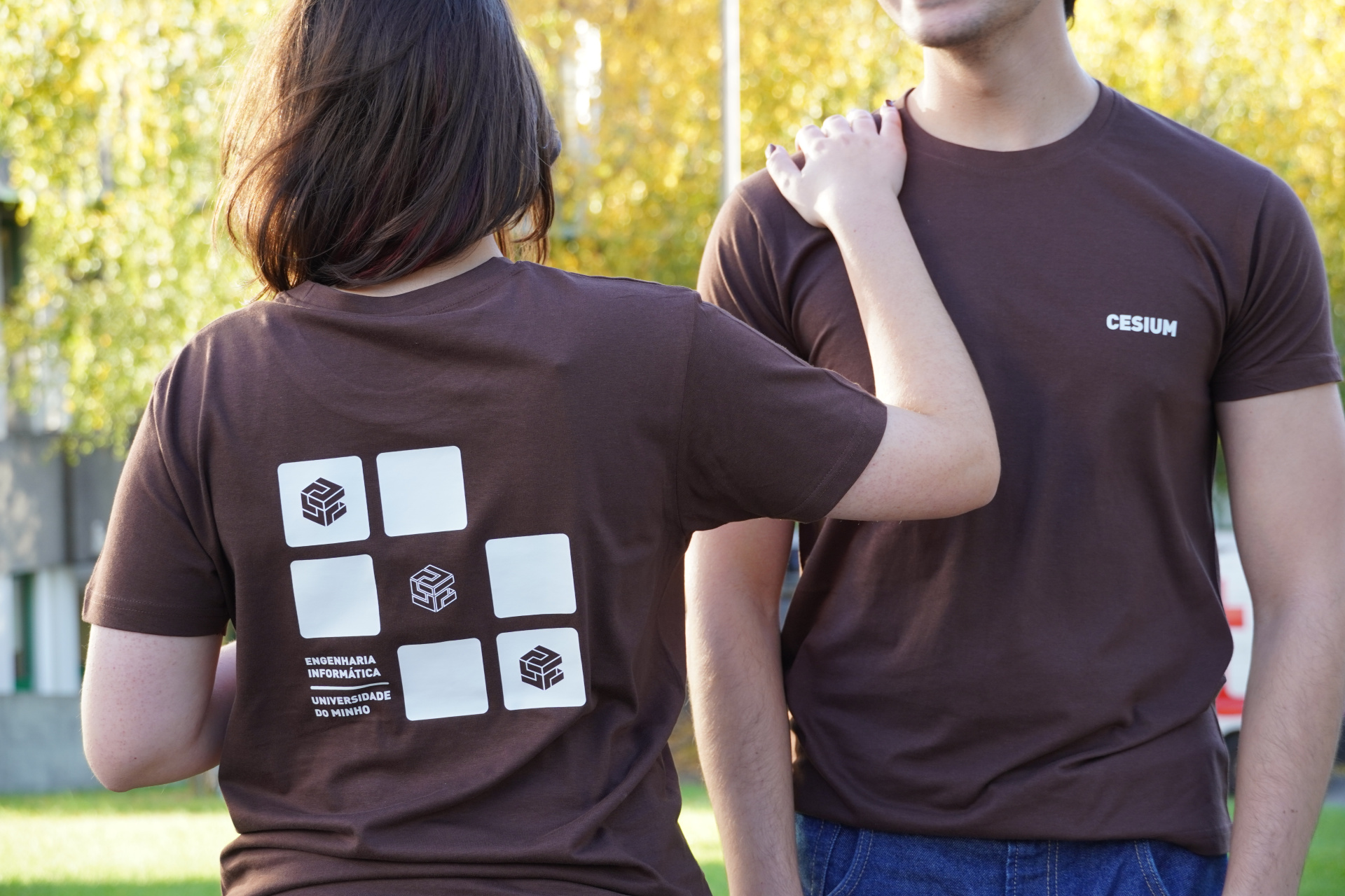 Par de t-shirts castanhas com estampados e textos relacionados à Universidade do Minho em ambiente exterior ensolarado