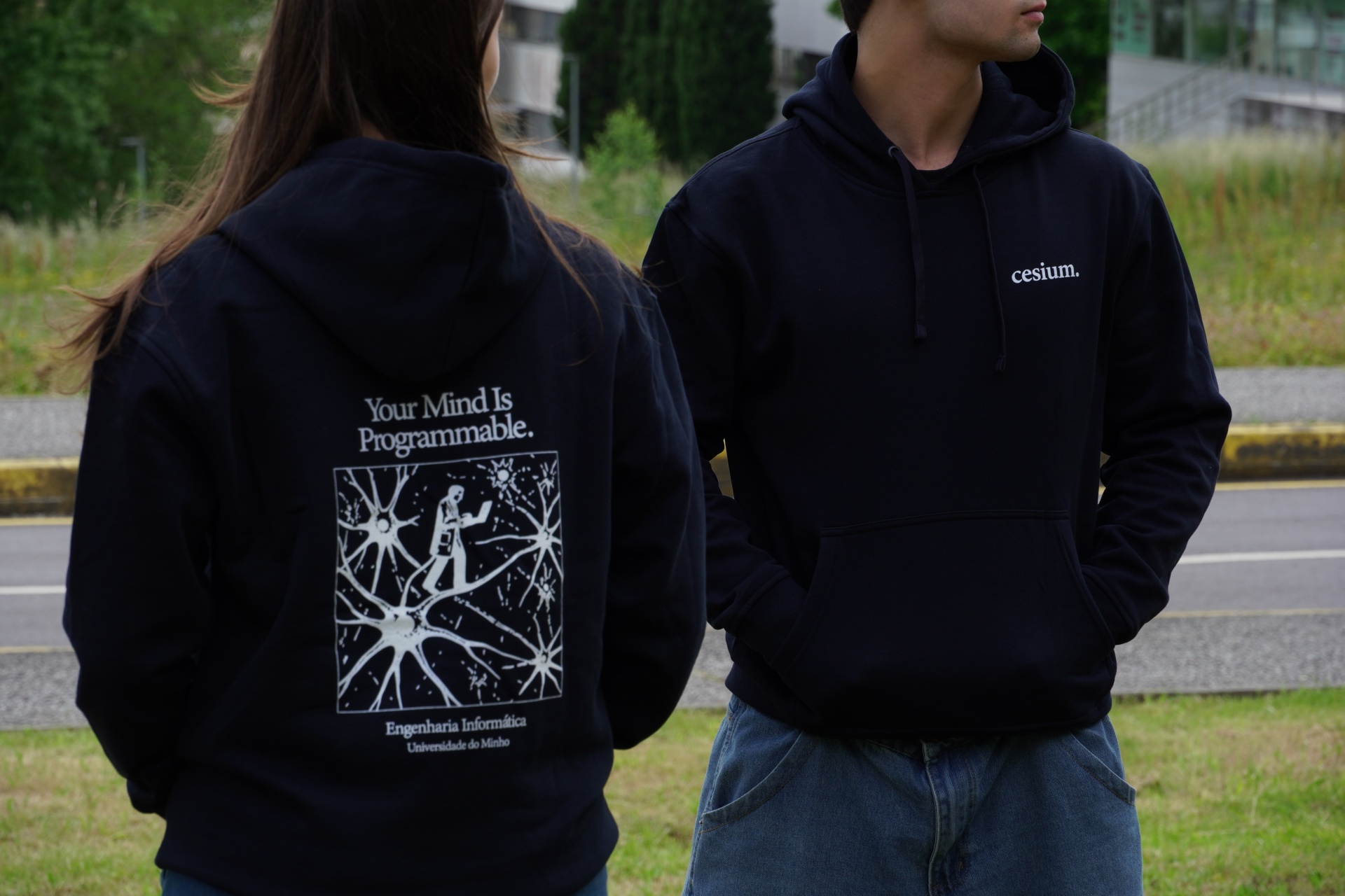 Dois hoodies pretos com textos e gráficos em branco, uma pessoa de costas e outra de frente ao ar livre