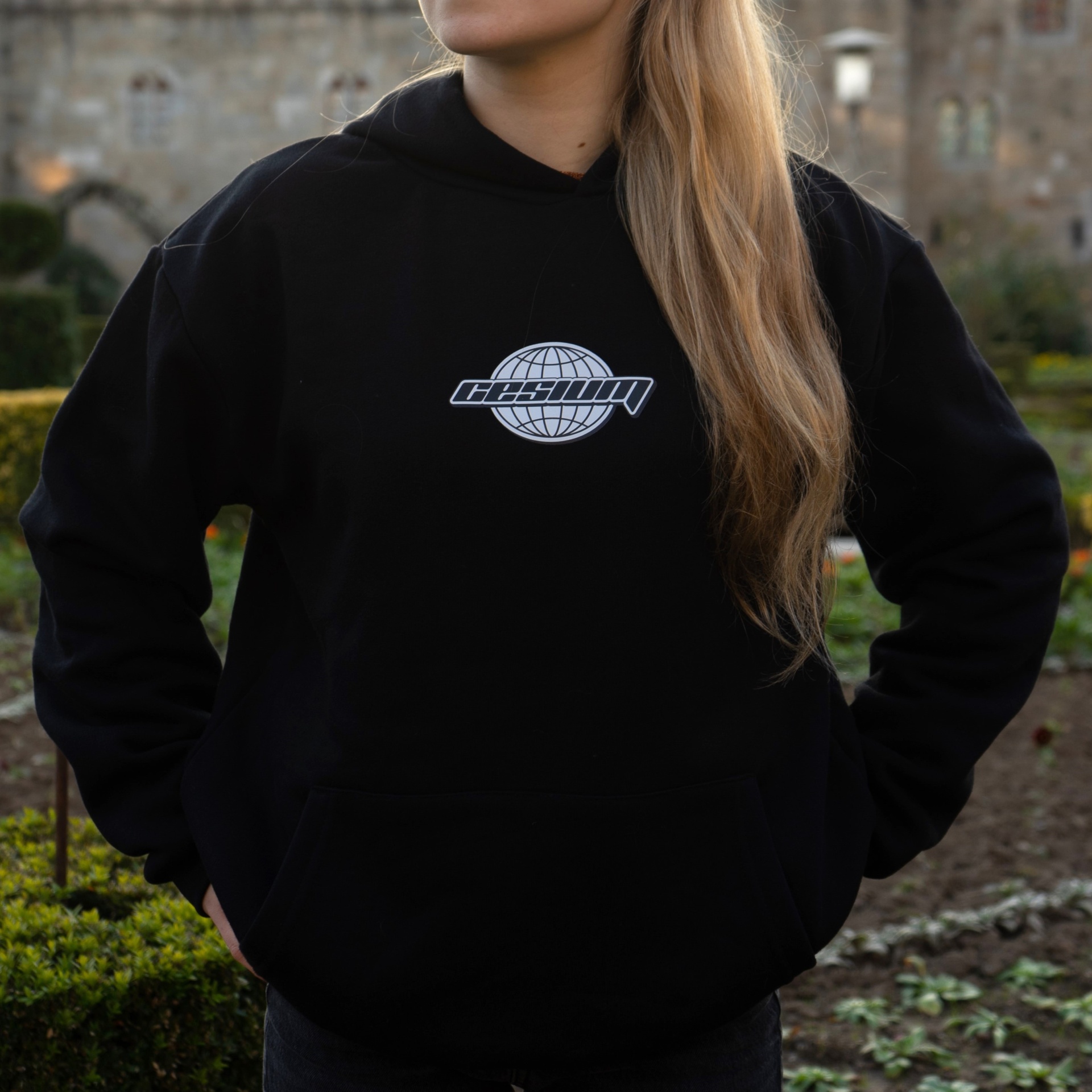 Sweatshirt preta com capuz e logotipo branco central, usada num jardim com plantas e edifícios ao fundo