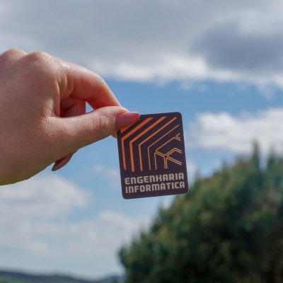 Cartão preto com linhas laranja e texto ENGENHARIA INFORMÁTICA segurado por mão em fundo de céu e árvores