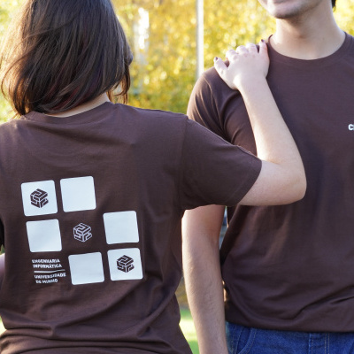 Par de t-shirts castanhas com estampados e textos relacionados à Universidade do Minho em ambiente exterior ensolarado