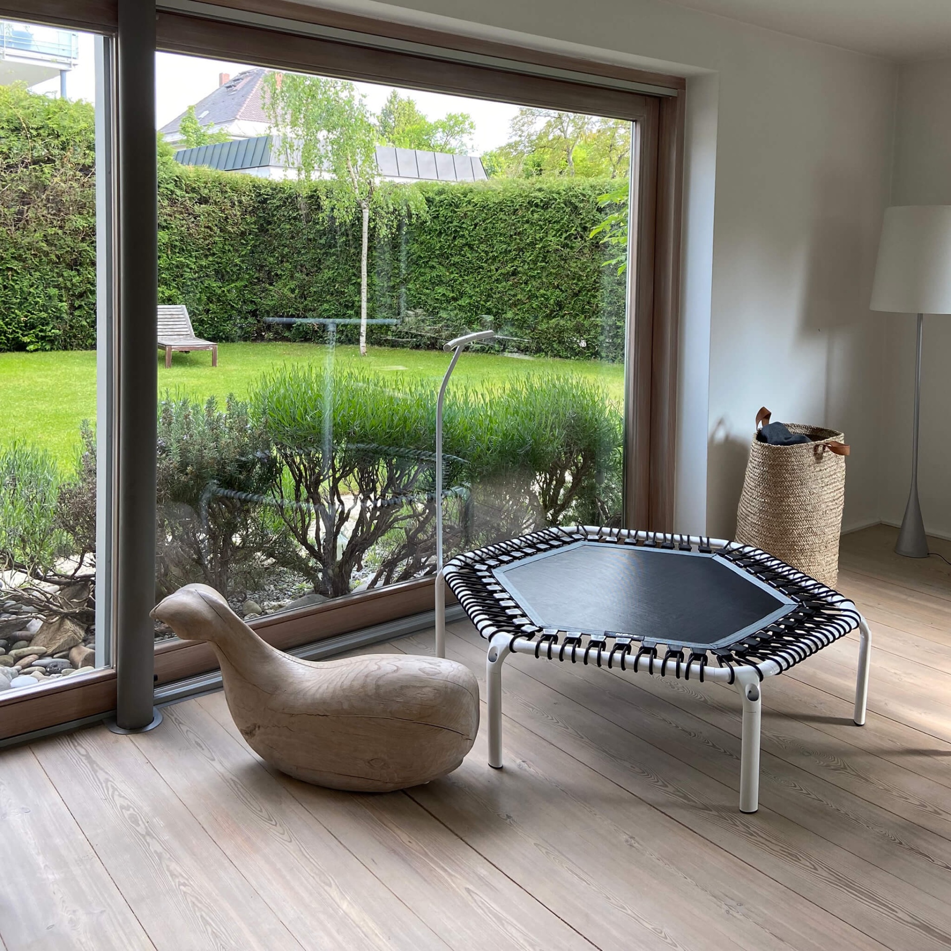 Sala com cadeira em madeira clara, mini trampolim preto, e cesta de ráfia junto a uma janela grande de madeira
