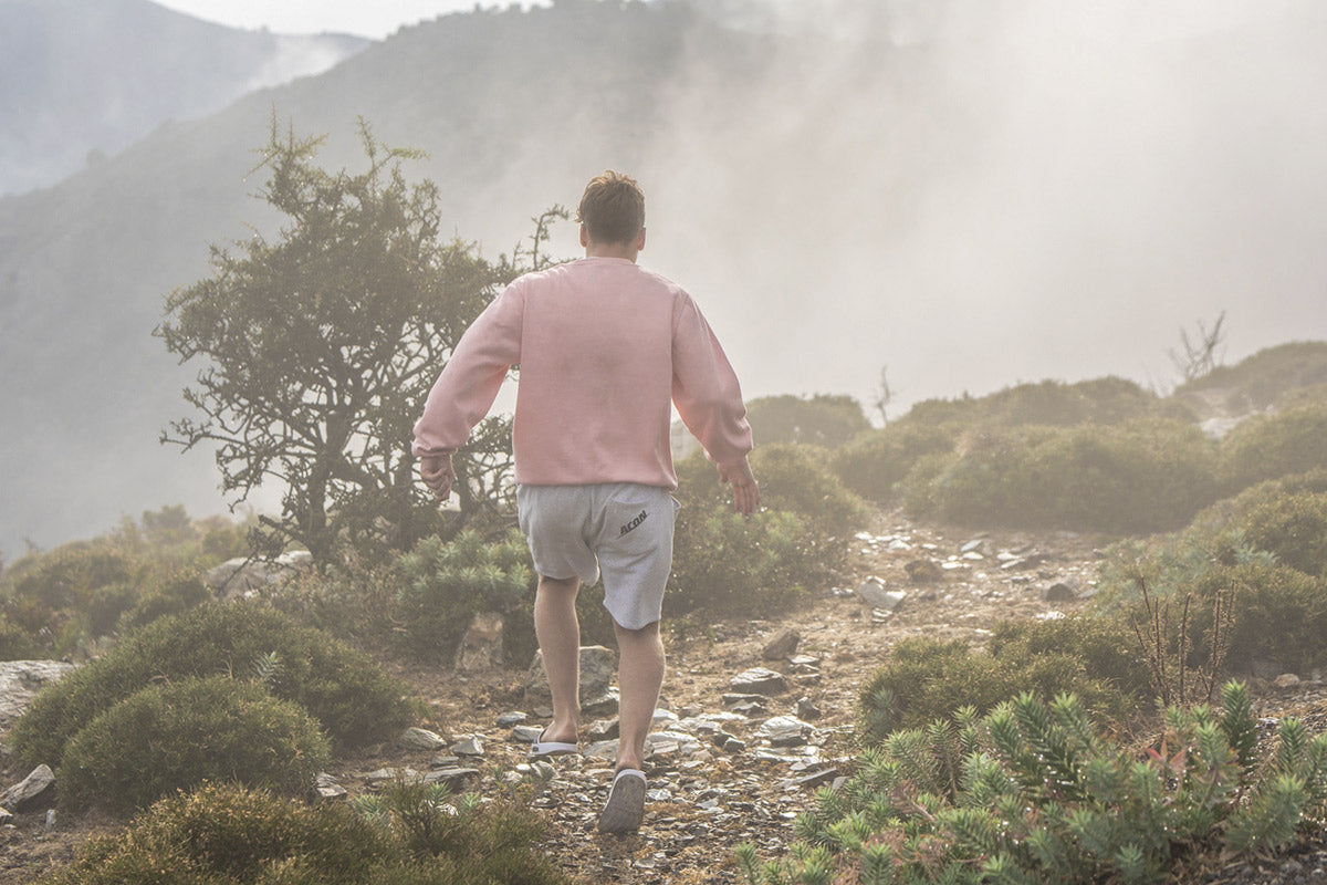 Homem de sweatshirt rosa e calções cinza a caminhar numa trilha de montanha