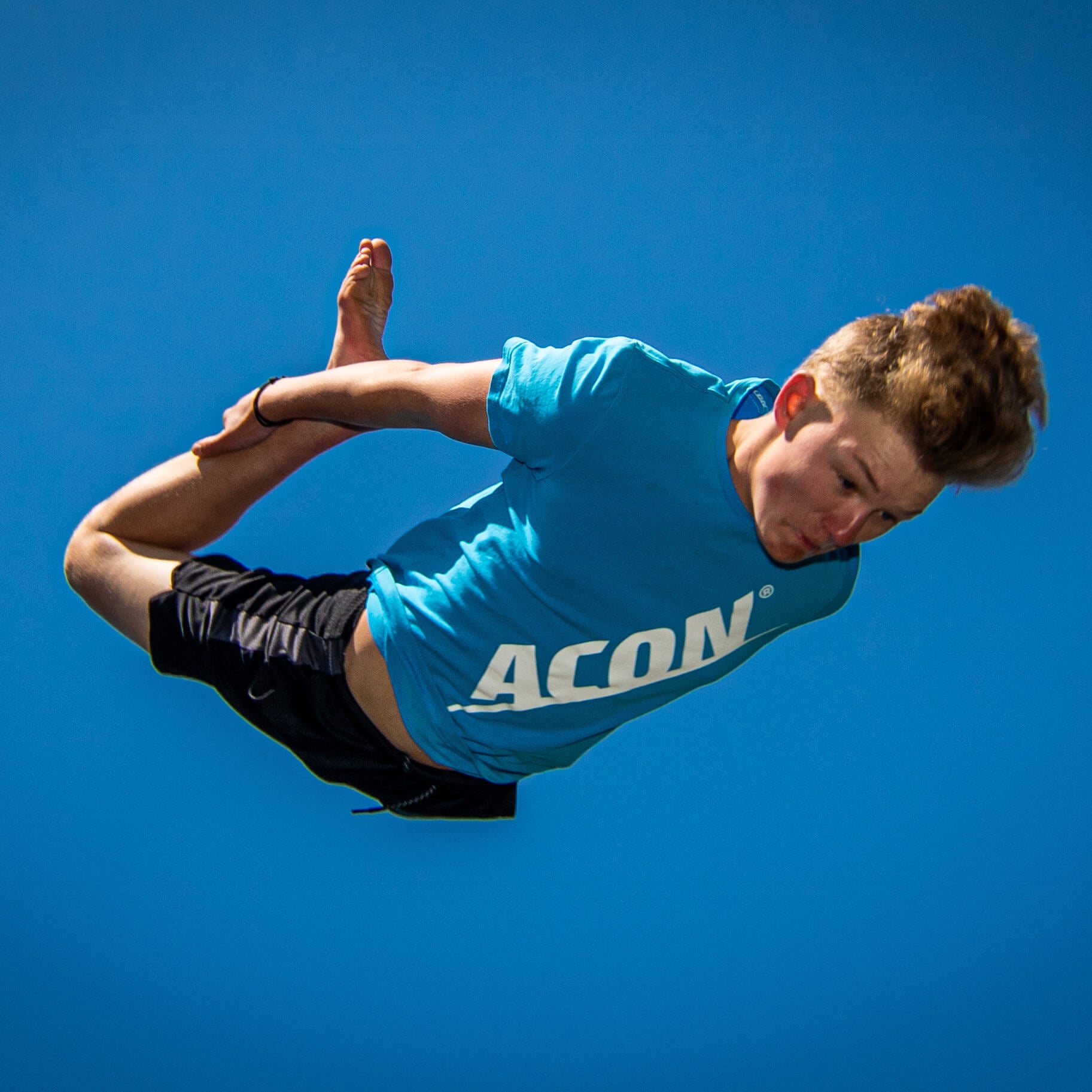 Jovem em pose acrobática vestindo t-shirt azul com texto branco e calções pretos, céu azul ao fundo