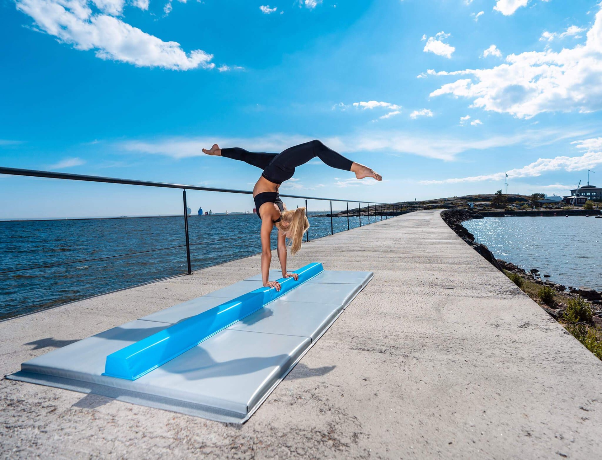 Mulher a fazer acrobacia sobre equipamento azul num tapete de ginástica cinza no passadiço junto ao mar