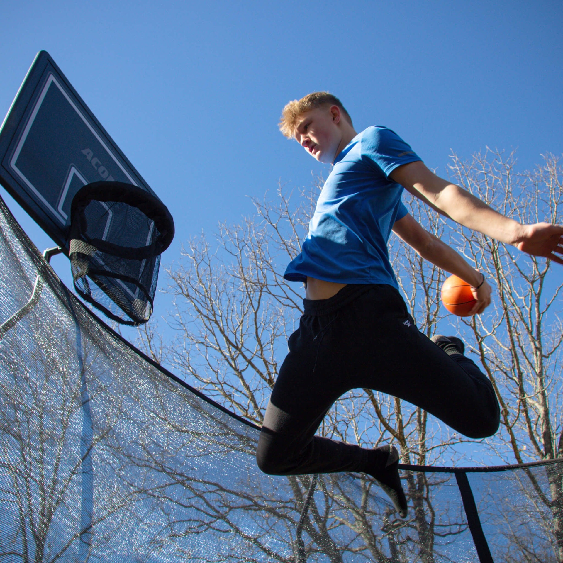 Jovem a saltar com bola de basquetebol junto a aro e rede preta num dia de céu azul.