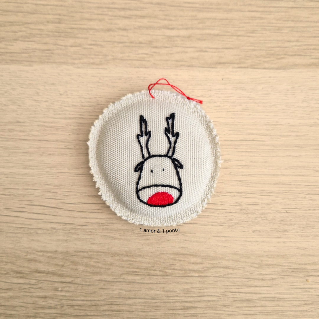 Pedaço redondo de tecido bege com bordas desfiadas e bordado de rena com nariz vermelho e chifres pretos