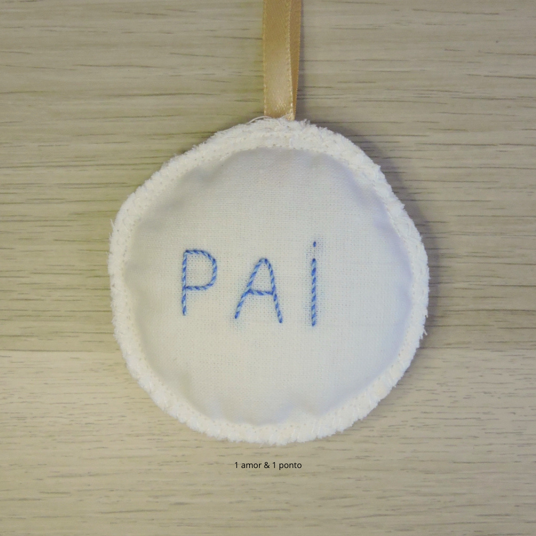 Pingente redondo de tecido branco com bordado azul 'PAI' e fita castanha