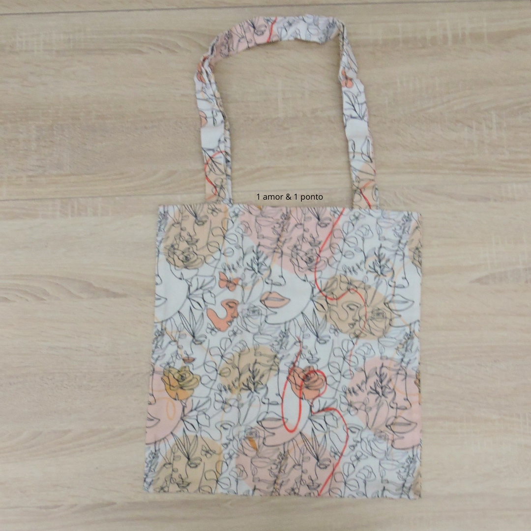 Saco de tecido com padrão artístico de faces femininas em cores pastel e texto '1 amor & 1 ponto'.