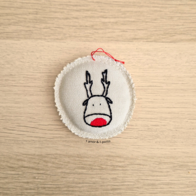 Pedaço redondo de tecido bege com bordas desfiadas e bordado de rena com nariz vermelho e chifres pretos