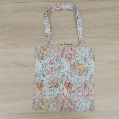 Saco de tecido com padrão artístico de faces femininas em cores pastel e texto '1 amor & 1 ponto'.