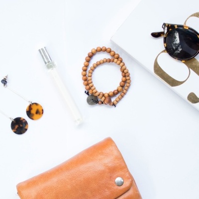 Artigos pessoais sobre fundo branco com sapatilhas padrão leopardo, brincos redondos castanho e preto, perfume, pulseira madeira, carteira castanha e óculos de sol.