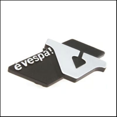 Emblema metálico preto e prateado com texto 'è vespa!' e letra V