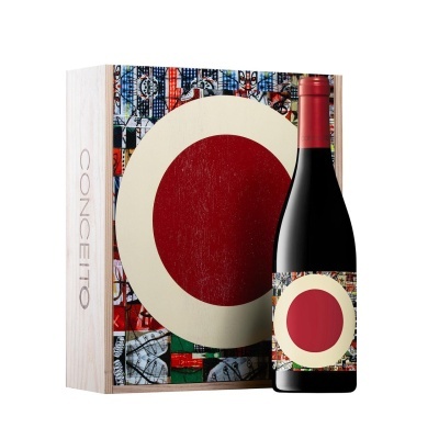 Garrafa de vinho tinto com rótulo vermelho, creme e colorido ao lado de caixa de madeira com a palavra CONCEITO