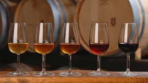 Cinco copos de vinho com várias cores em cima de mesa de madeira com barris ao fundo