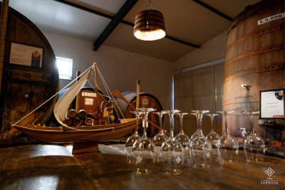 Adega com barris de madeira, miniatura de barco, copos de vinho e certificado emoldurado.