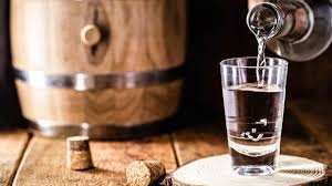 Copo de vidro sendo enchido com líquido claro ao lado de rolhas e barril de madeira.