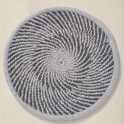 Tapete redondo tecido à mão em tons de cinza com padrão espiral sobre tecido bege.