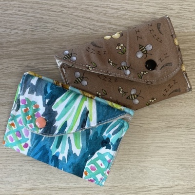 Dois porta-moedas de tecido com padrões coloridos, um com abelhas e outro com flores, sobre mesa de madeira clara.