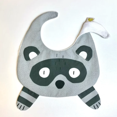 Babete infantil em tecido cinzento com desenho de guaxinim e etiqueta amarela