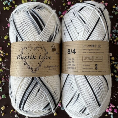 Dois novelos de fio Rustik Love branco com riscas pretas, rótulos castanhos e fundo com flores coloridas.