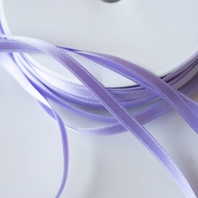 Rolo de fita de cetim lilás desenrolado sobre superfície branca