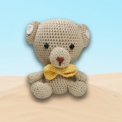 Ursinho de croché bege com laço amarelo sentado sobre superfície clara